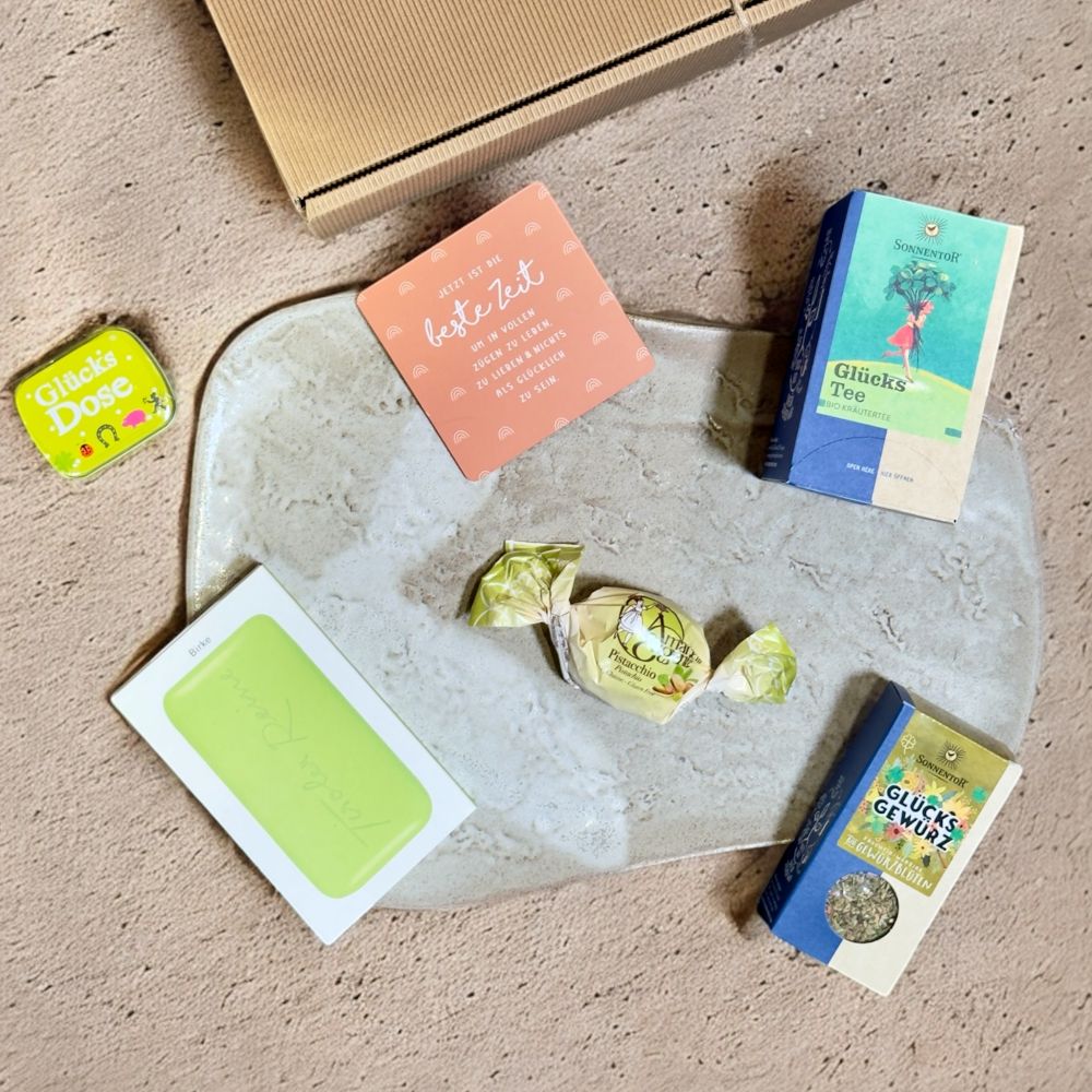 Ausgepackte Glücks Geschenkbox mit Sonnentor Glücks Tee Sonnentor Glücks Gewürzblüten Tiroler Reine Seife Birke das Glück Amaretti Eugenia GRUSS CO Mintdose Glücksdose und Grafik Werkstatt Korkuntersetzer Jetzt ist die beste Zeit